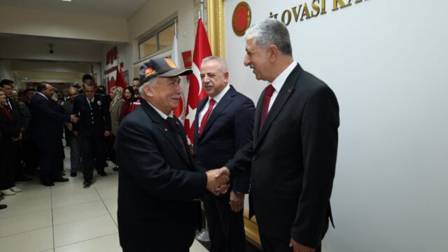 Kaymakamlık binasında şapkalı yaşlı bir adam ve takım elbiseli iki adam tokalaşıyor; arkada Türk bayrakları var.