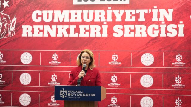 Kırmızı ceketli bir kadın, sergi panosunun önündeki kürsüde mikrofonla konuşuyor.