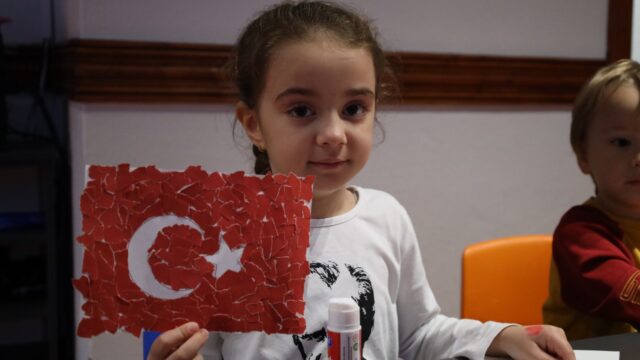 Küçük bir kız, kırmızı kâğıt parçalarından yaptığı Türk bayrağı kolajını gösteriyor; arkada bir çocuk daha var.