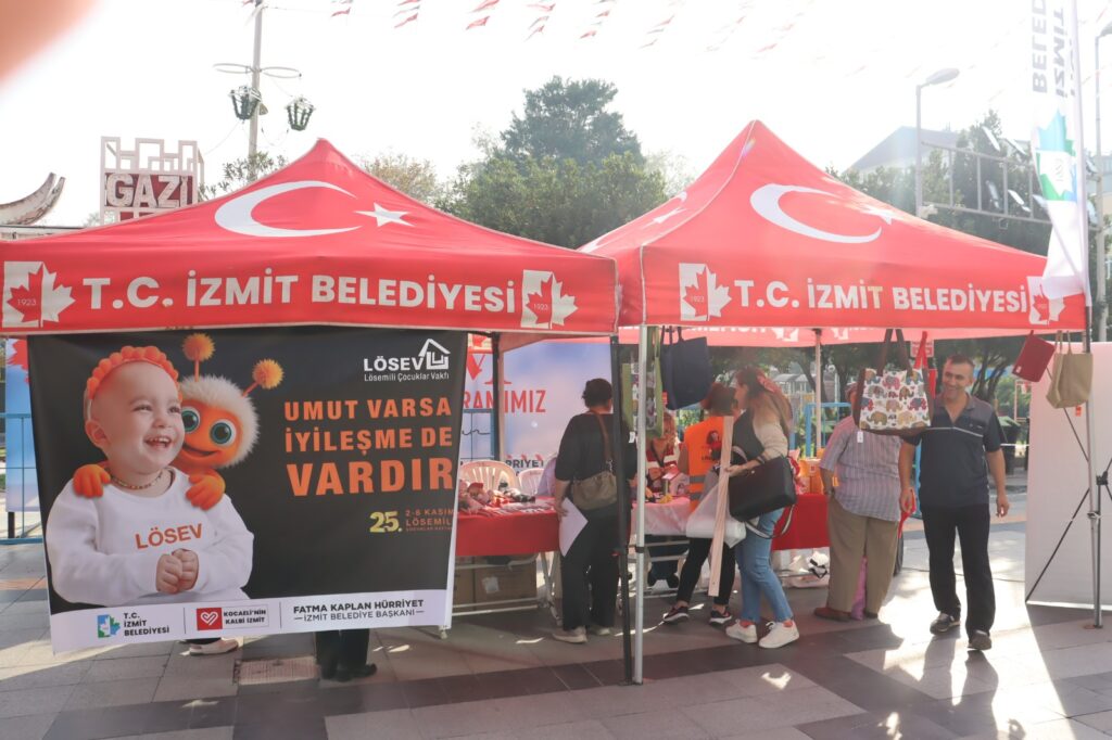 İzmit Belediyesi’nin kırmızı çadırlarında LÖSEV standı; önde gülen bebekli afiş, çadırın altında alışveriş yapan insanlar.