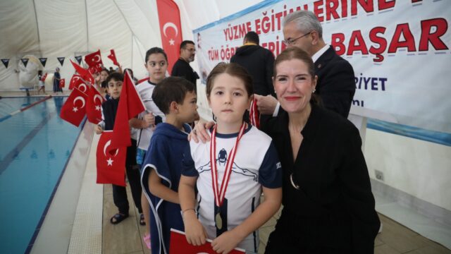 Boynunda madalya olan bir kız çocuğu, yanında siyah giyimli bir kadın ve Türk bayraklı çocuklarla havuz kenarında.