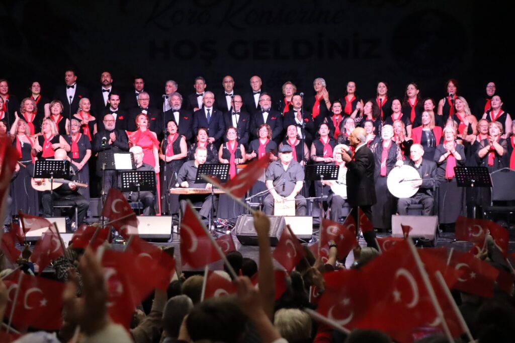 Sahnede koro ve saz sanatçıları, önde Türk bayrağı sallayan seyirciler