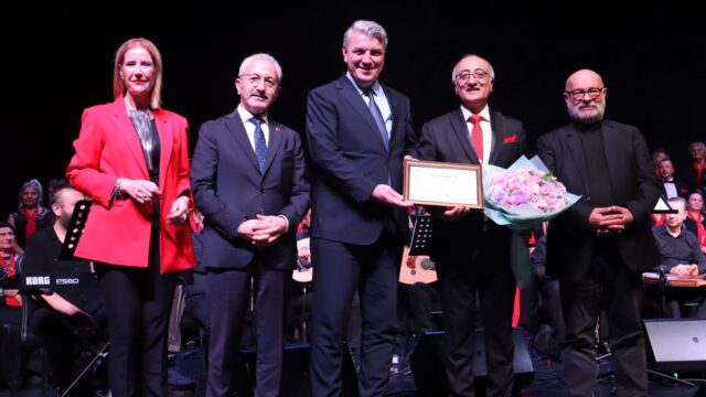 Sahnede beş kişi poz veriyor; biri çiçek ve çerçeveli belge tutuyor