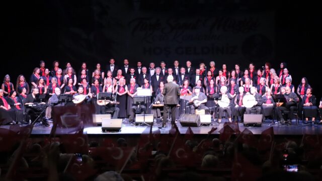 Geniş sahnede koro ve orkestra, önde bayrak sallayan seyirci silüetleri