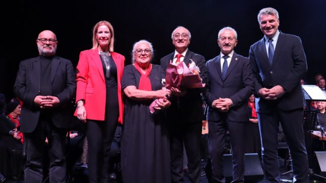 Sahnede altı kişi çiçekle poz veriyor; kırmızı ceketli bir kadın ve takım elbiseli erkekler var