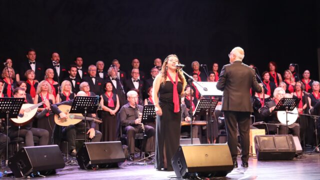 Kırmızı fularlı kadın solist mikrofonda şarkı söylüyor, arkada koro ve sazlar