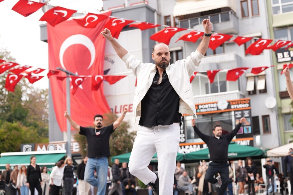 Beyaz ceketli bir adam havada zeybek figürü yapıyor, arkasında dans eden iki kişi ve büyük bir Türk bayrağı var.