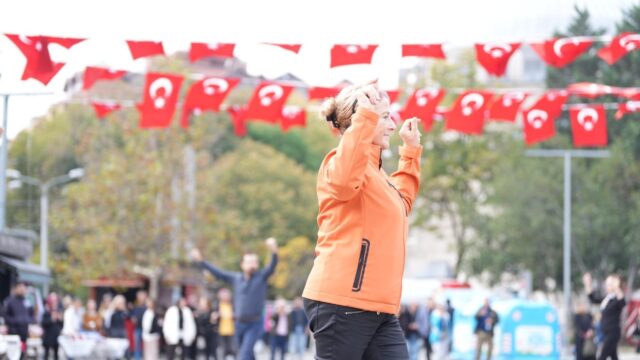 Turuncu ceketli bir kadın gülümseyerek kollarını kaldırıp dans ediyor, arkada Türk bayrakları ve izleyiciler var.