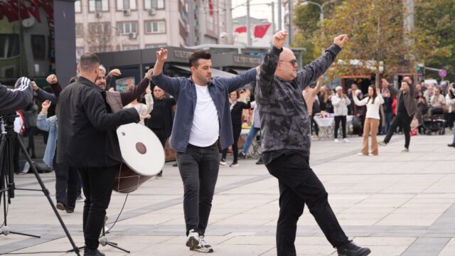 İki erkek kollarını açarak zeybek oynuyor, solda davul çalan bir kişi ve çevrede izleyiciler var.