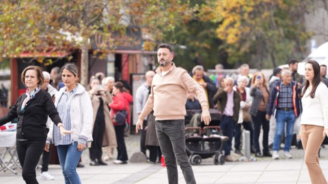Açık renk kazaklı sakallı bir adam ve çevresindeki kadınlar meydanda dans figürü yapıyor, arkada izleyiciler var.