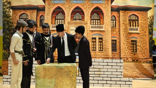 Öğrenciler tarihî kıyafetlerle sahnede harita masasının başında oyun oynuyor; arkada taş bina dekoru var.