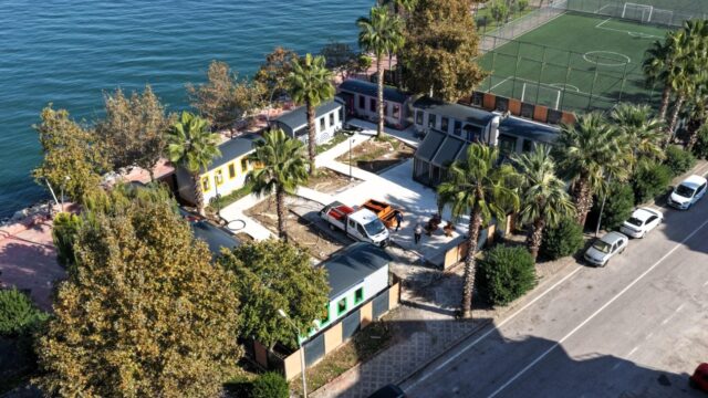 Sahil boyunca palmiyeler arasında renkli küçük yapılar, yanında halı saha ve cadde, havadan görünüm.