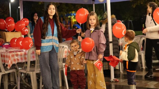 Mavi yelekli genç bir görevli ve çocuklar, kırmızı balonlar ile Türk bayrakları tutarak çadırın önünde duruyor.