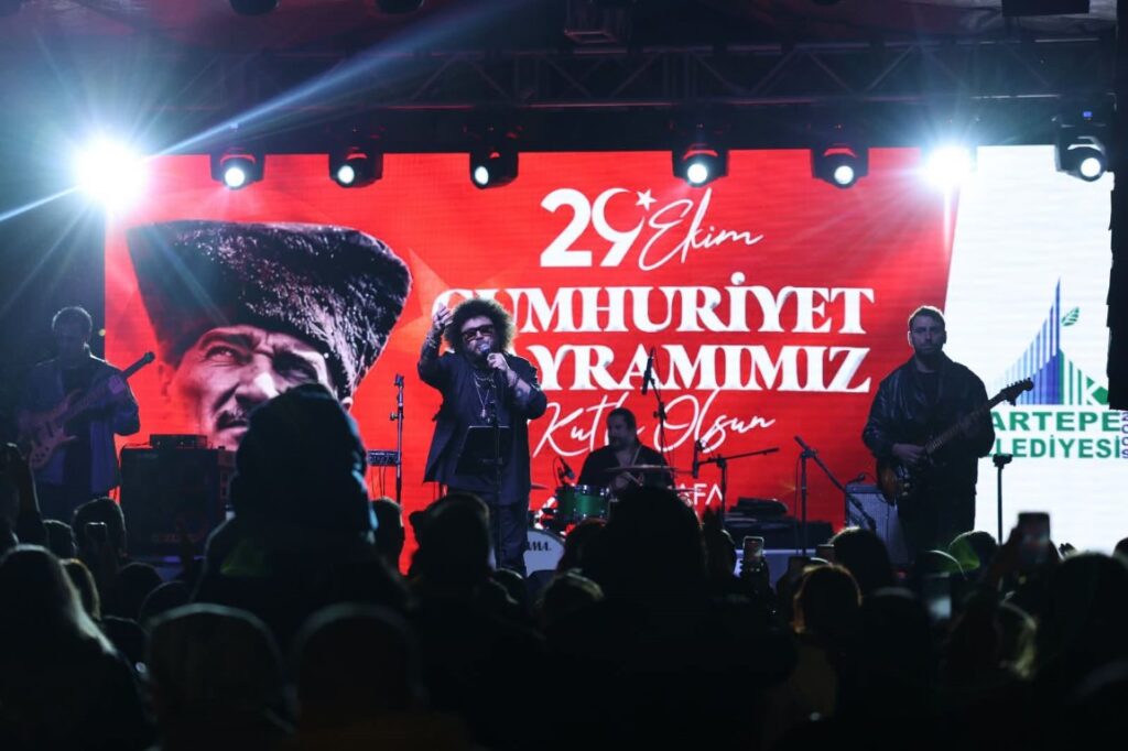 Sahnede grup konser veriyor; arkadaki ekranda Atatürk görseli ve 29 Ekim Cumhuriyet Bayramı yazısı var.