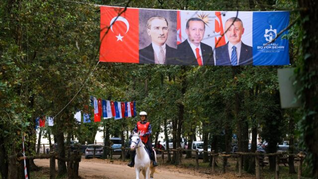 Kırmızı yelekli ve kasklı binici beyaz atla ormanlık yolda ilerliyor; yukarıda üç portreli büyük bir pano asılı.
