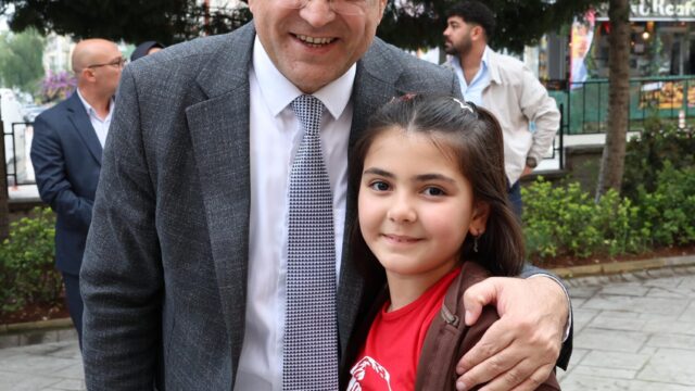Gözlüklü, gri ceketli bir adam kırmızı tişörtlü bir kız çocuğuyla poz veriyor; arkada ağaçlar ve yürüyen insanlar var.