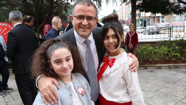 Gözlüklü, takım elbiseli bir adam iki kız çocuğunun omzuna sarılmış, gülümsüyor; arkada kalabalık ve çelenk var.