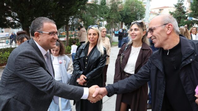 Takım elbiseli bir adam, açık havada yaşlı bir erkekle el sıkışıyor; çevrelerinde gülümseyen kadınlar var.