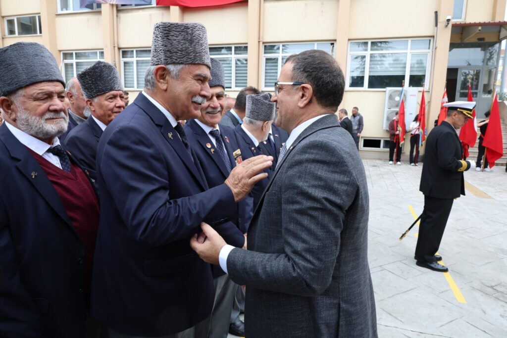 Takım elbiseli bir adam, kalpaklı yaşlı gazilerle selamlaşıyor; arkada bayraklı bir tören alanı var.
