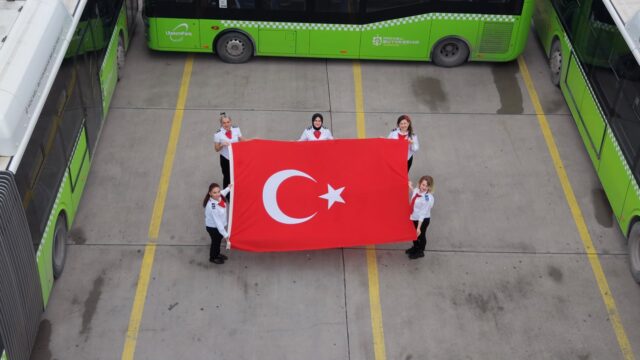 Beş kadın çalışan yeşil otobüslerin önünde büyük bir Türk bayrağını açmış tutuyor, yukarıdan çekim