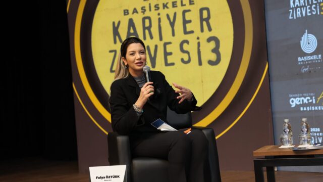 Sahnede siyah koltukta oturan, elinde mikrofonla konuşan siyah giysili kadın moderatör; arkada zirve panosu var.