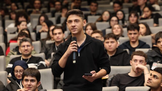 Kalabalık salonda ayakta mikrofonla konuşan siyah kazaklı genç, çevresinde dinleyiciler oturuyor.