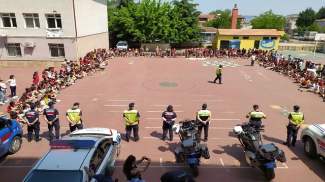 Okul bahçesinde jandarma ve trafik ekipleri, motosiklet ve araçlarla sıralanmış; çevrede oturan öğrenciler izliyor.