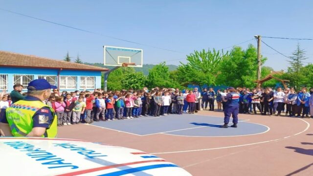 Okul bahçesindeki basketbol sahasında öğrenciler toplanmış, bir jandarma personeli karşılarında konuşuyor; önde jandarma aracı görünüyor.