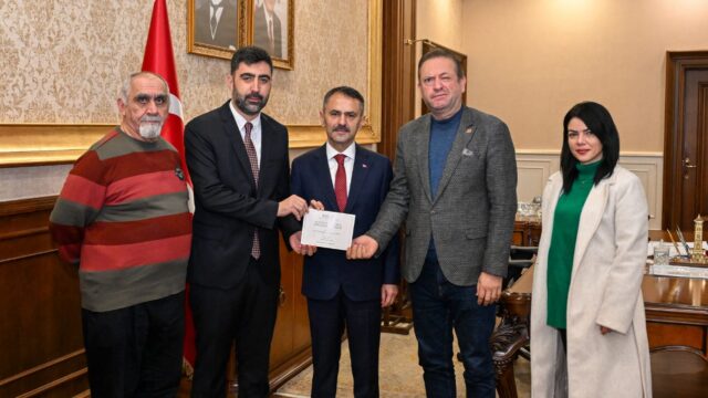 Makam odasında bayrağın yanında dört erkek ve bir kadın poz veriyor; ortadaki takım elbiseli adama zarf uzatılıyor.