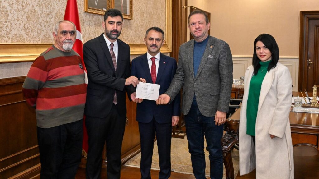 Makam odasında bayrağın yanında dört erkek ve bir kadın poz veriyor; ortadaki takım elbiseli adama zarf uzatılıyor.