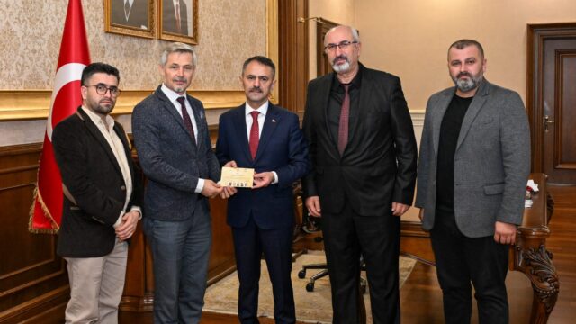 Makam odasında, Türk bayrağı ve portrelerin önünde beş erkek bir belge zarfı tutarak poz veriyor.