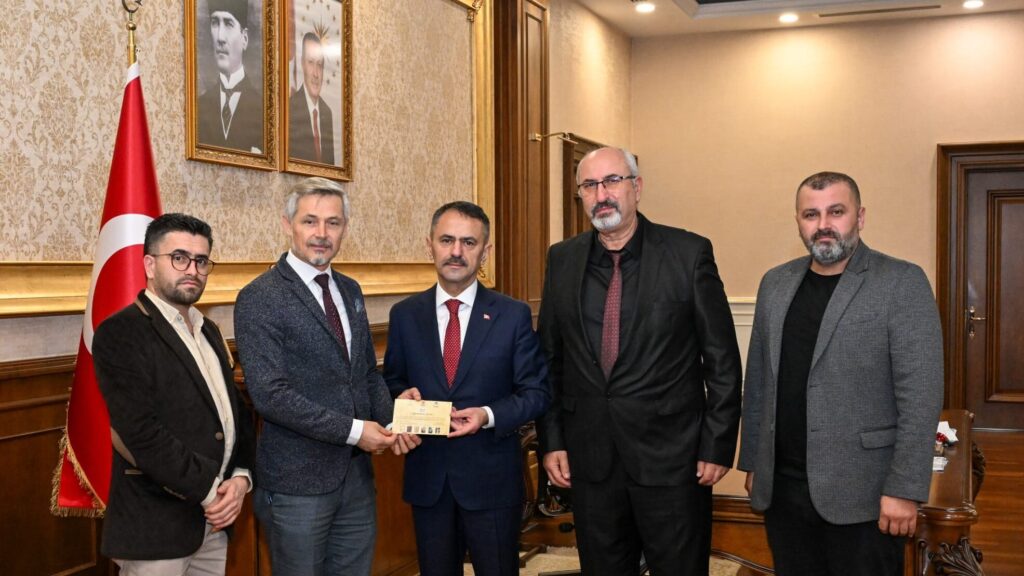 Makam odasında, Türk bayrağı ve portrelerin önünde beş erkek bir belge zarfı tutarak poz veriyor.