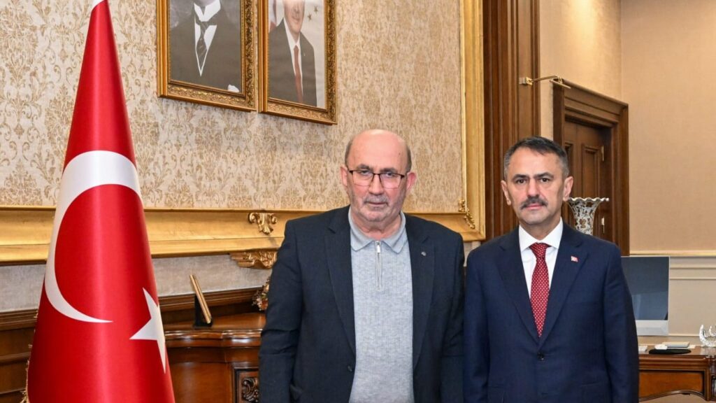 Makam odasında, Türk bayrağı ve iki portrenin önünde koyu takım elbiseli iki erkek yan yana poz veriyor.