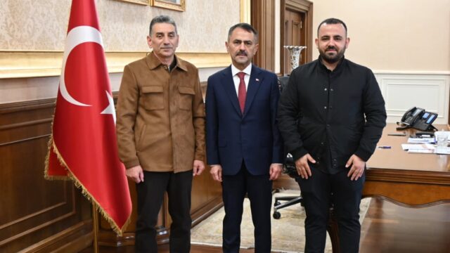 Makam odasında Türk bayrağının yanında kahverengi ceketli, takım elbiseli ve siyah giyimli üç erkek kameraya poz veriyor.