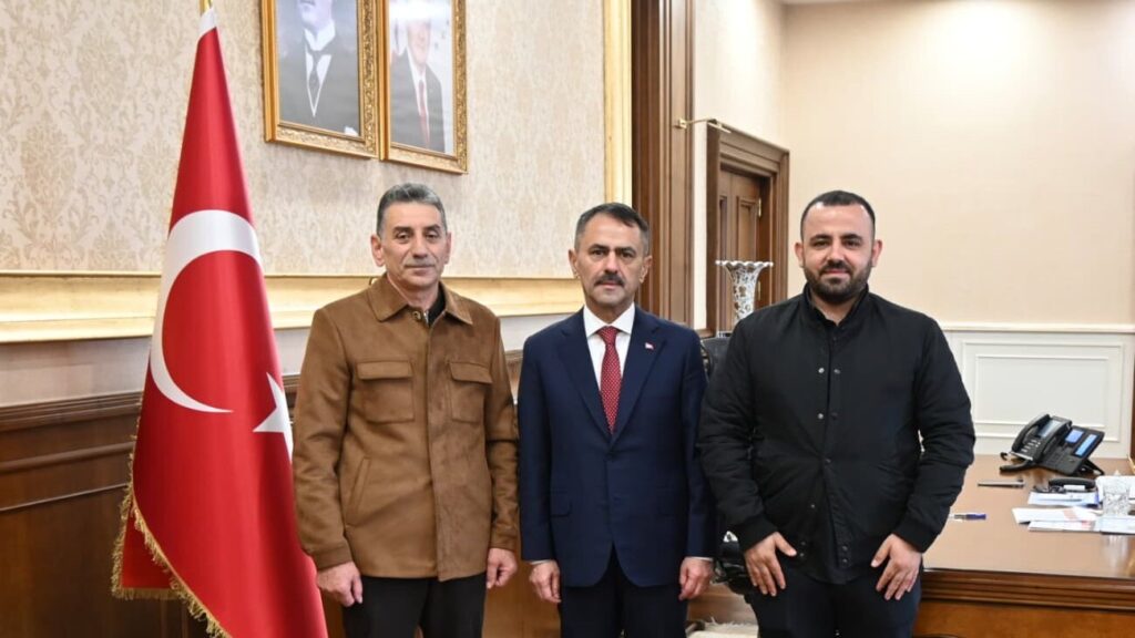 Makam odasında Türk bayrağının yanında kahverengi ceketli, takım elbiseli ve siyah giyimli üç erkek kameraya poz veriyor.