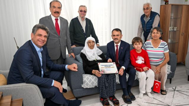 Salonda gri kanepede oturan beyaz başörtülü yaşlı kadın belgeyi tutuyor; yanında erkekler, bir kadın ve kırmızı tişörtlü çocuk var.