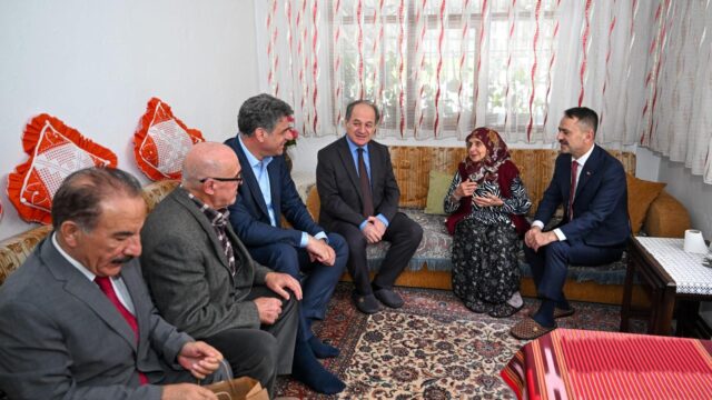 Evde koltukta oturan başörtülü yaşlı kadın, yanında takım elbiseli dört erkek ve ön planda iki erkek sohbet ediyor.