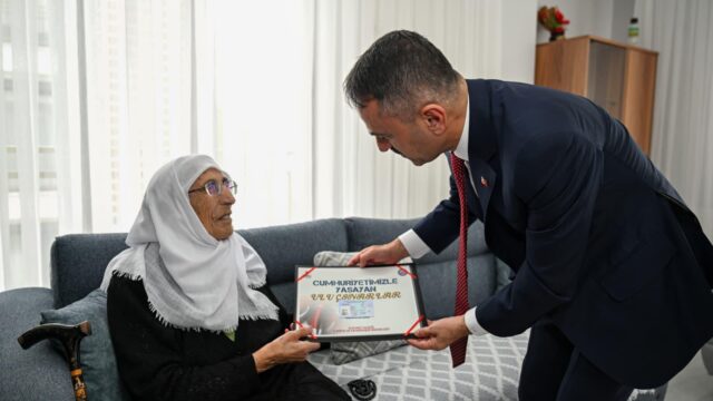 Gözlüklü, beyaz başörtülü yaşlı kadın, öne eğilen takım elbiseli bir erkekle birlikte çerçeveli Ulu Çınarlar belgesini tutuyor.