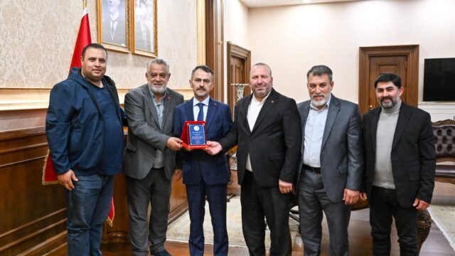 Makam odasında altı erkek yan yana duruyor; ortadaki lacivert takımlı kişi elinde mavi-kırmızı bir plaket tutuyor.