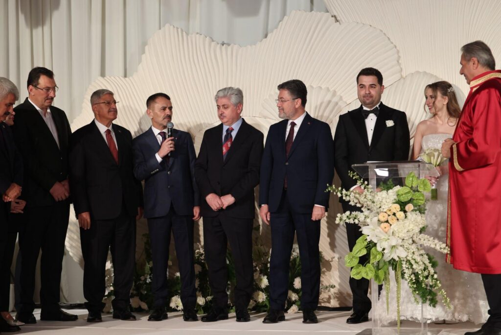Takım elbiseli erkekler sahnede sıra halinde duruyor; biri mikrofonla konuşuyor, yanlarında damat, gelin ve kırmızı cübbeli görevli var.